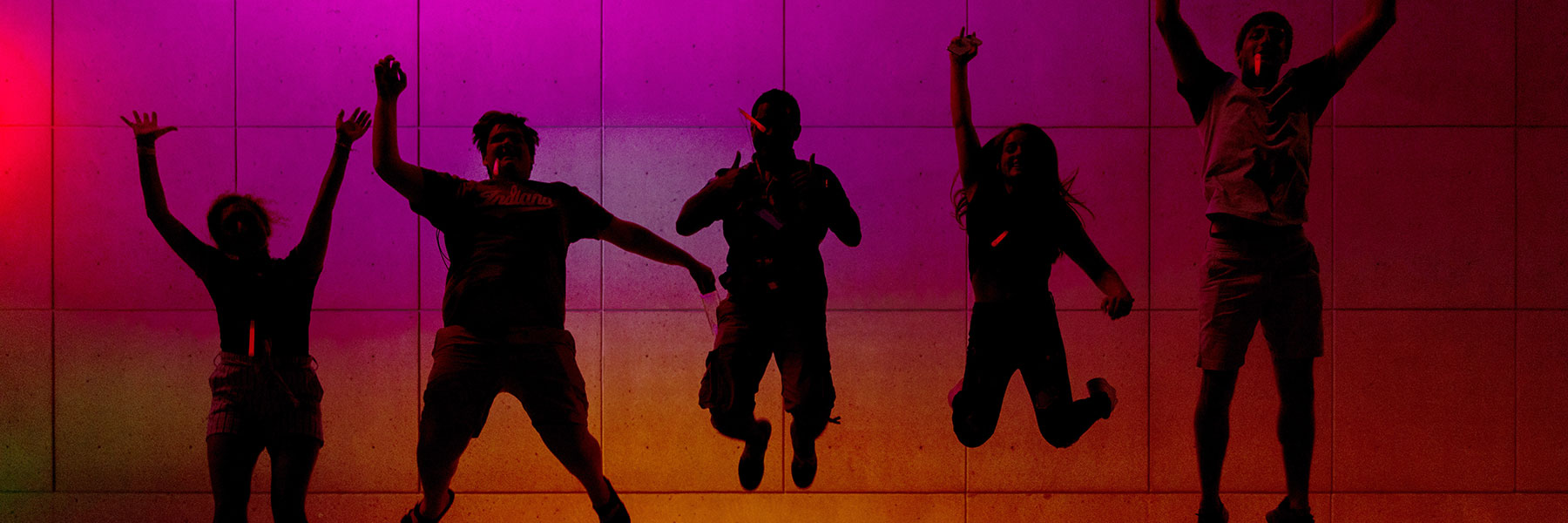 Students jumping in the air at night in front of colored lights.
